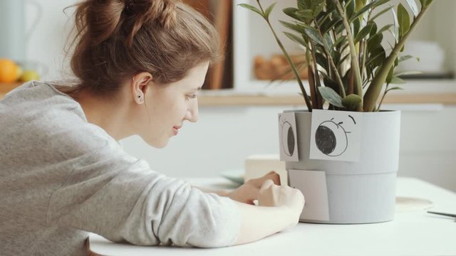 Side view of young positive woman sitting at table and drawing smiley face on sticky notes attached to plant pot and enjoying it staying alone at home on quarantine