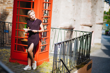 A girl with a book is standing by the red telephone box.