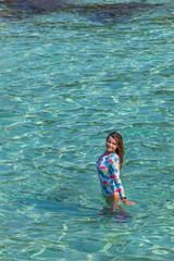 A beautiful girl in a one-piece swimsuit stands in the clear, turquoise water and looks up. Photo from the top