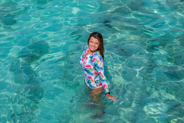 A beautiful girl in a one-piece swimsuit stands in the clear, turquoise water and looks up. Photo from the top