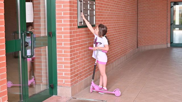 A little girl calls the intercom to go home. The child wants to get into the house. Girl 7 years old with brown hair and a scooter.
