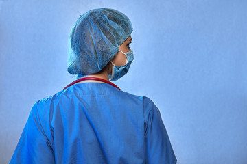 Doctor from the back in medical uniform, protective mask, surgical cap, with stethoscope isolated on blue background. Coronavirus Epidemic COVID-19, space for text