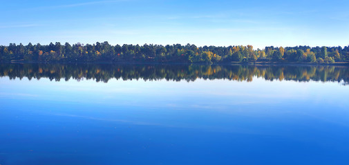 The blue sky is reflected in the clear water of the lake against the background of the forest in the distance. 