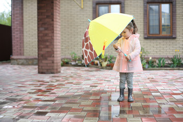The rain has just ended. Little girl standing under a children's umbrella.  The girl is wearing rubber boots and a raincoat.