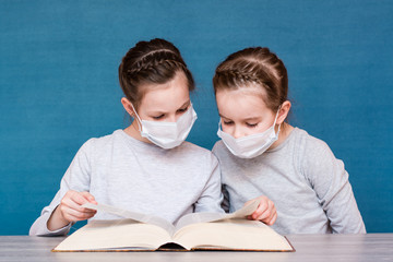 Girls in medical masks in quarantine carefully read a book at the table. Education children in...