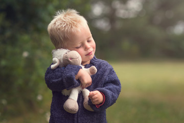 Little blond boy holding plush sheep in the nature
