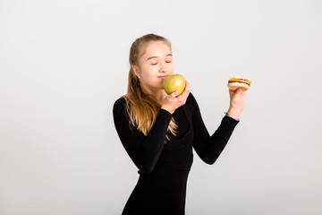 young slender girl holds a hamburger and an apple against a white background. choosing healthy...
