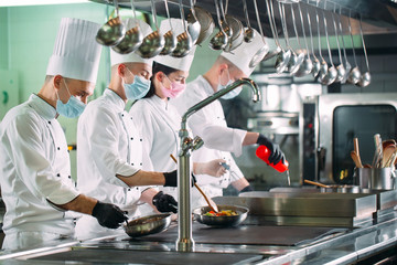 Chefs in protective masks and gloves prepare food in the kitchen of a restaurant or hotel.