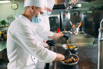 Chefs in protective masks and gloves prepare food in the kitchen of a restaurant or hotel.