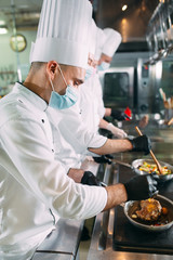 Chefs in protective masks and gloves prepare food in the kitchen of a restaurant or hotel.