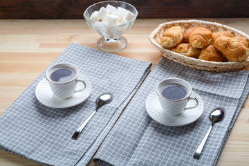 Two cups of coffee on napkins on a wooden table with croissants and sugar.
