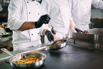 Chefs in protective masks and gloves prepare food in the kitchen of a restaurant or hotel.
