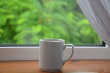 A white cup of tea or coffee stands on a wooden window sill against the background of a summer garden