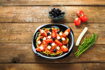 Black plate with spaghetti in a sauce decorated with cherry tomatoes olives and mozzarella on a wooden table.