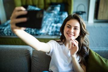 Portrait of a happy young woman taking a selfie with mobile phone while leaning on a couch at home