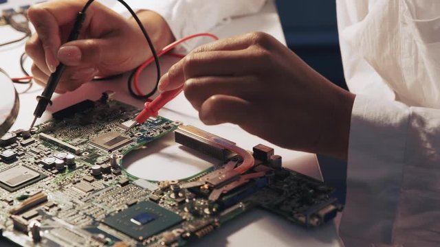 Close up of engineer's hands using multimeter tester to check motherboard in laboratory. The specialist is in white robe