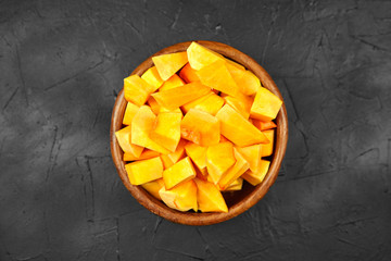 Pumpkin pieces in a round wooden bowl on black table, top view. Chopped pumpkin (butternut squash)...