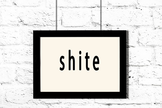 Black frame hanging on white brick wall with inscription shite