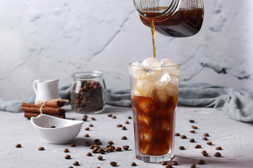 Cold coffee is poured into a tall glass with ice cubes on light gray background, Horizontal format