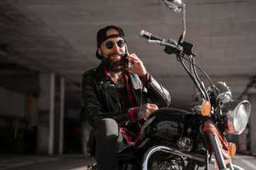 Plakat Smiling charming attractive bearded man biker on motorcycle calling mobile cell phone, wearing black and sunglasses, freedom concept. 