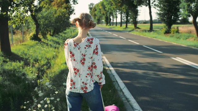 Young woman hitchhiking on countryside road. Back view of young female with red suitcase standing on roadside and hitchhiking while travelling through countryside.