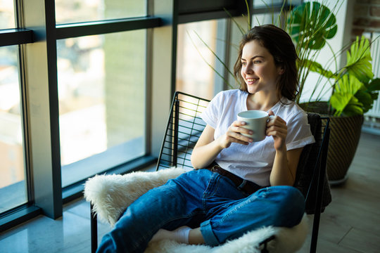 Young woman at home sitting on modern chair in front of window relaxing in her living room drinking coffee or tea