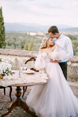 Wedding at an old winery villa in Tuscany, Italy. A wedding couple is standing near the table for a wedding dinner, the groom hugs the bride by the waist.