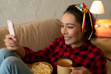 Happy cheerful Young brazilian woman with tea mug watching video on mobile smart phone Indoors at home living room. Entertainment, recreation, live stream concept.