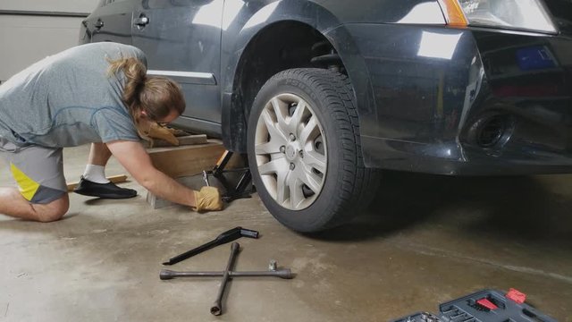 Time lapse of a man in glasses and ponytail wearing shorts use hand jack to rise passenger car while sliding wood logs to stabilize car for later repair or maintenance.