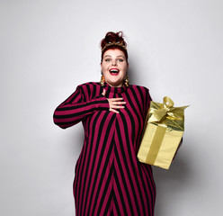 Big ginger woman in striped dress, crown and earrings. Holding golden gift box, looking joyful,...