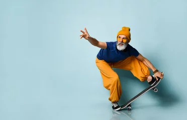 Fototapeten Gray-bearded grandpa in t-shirt, sunglasses, orange pants, hat, gumshoes. Riding black skateboard, posing on blue background © FAB.1