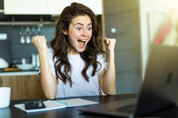 Excited young woman with win gesture with fist up sitting at table and looking at laptop