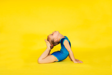 Young cute barefooted gymnast girl with two buns in blue sport suit posing on yellow background with copy space.