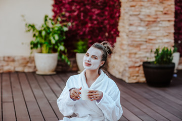 Young pretty woman in white bathrobe and mask on her face posing outdoor with cup of coffee