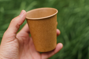 Close up isolated eco-friendly natural coffee cup. Disposable ecological utensils in hands on green grass background. Sustainability of planet. Cardboard glass made of fiber of bamboo and bagasse