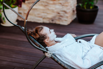 Young pretty woman in white bathrobe and mask on her face posing outdoor on sun bed