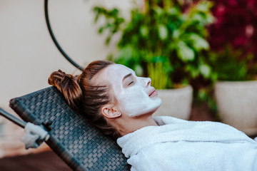 Young pretty woman in white bathrobe and mask on her face posing outdoor on sun bed
