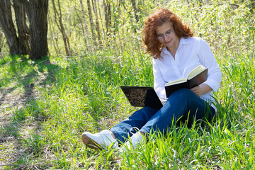 Portrait of a red-haired girl in glasses working outdoors. Makes notes in the diary. On the grass lies a laptop