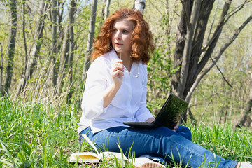 Obraz premium The girl with red hair thought holding glasses in her hands. Online work in nature. Grass, trees.