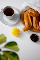Fresh crispy rolls under white kitchen towel on wooden cutting board, cup of black tea on plate, jam and honey on white table for breakfast