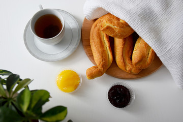 Fresh crispy buns under white kitchen napkin on wooden cutting board, jam, honey, cup of hot tea served for breakfast on white table