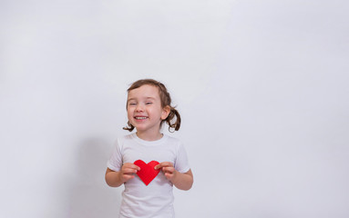 A little girl in a white t-shirt smiles and holds a small red cardboard heart on a white background with space for text.