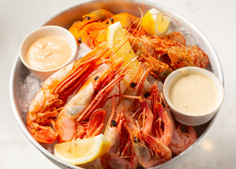 Freshly cooked assorted shrimps on the ice plate with lemon slices and sauces on a stone countertop.