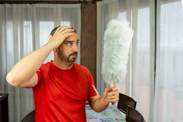 Man with a beard and short hair in a red shirt with a feather duster to clean the pole in the living room at home
