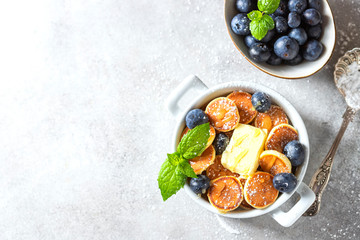 Bowl with tiny pancake cereal on a light concrete background. Trendy food. Mini cereal pancakes with butter, honey and blueberries.