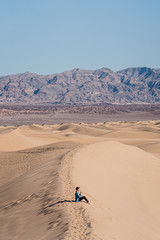 A travel girl is sitting on the top of a sand dune back to the camera. A traveler in a black hat and blue skirt in Death Valley National Park. A small figure of a person around nature