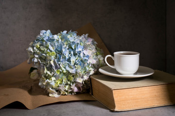 A branch of green pastel hydrangeain with white cup of coffee and vintage book on a on a gray craft vintage background.