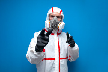 man in a chemical protective suit shows his fingers at you on a blue background, an epidemiologist, biologist, virologist, scientist