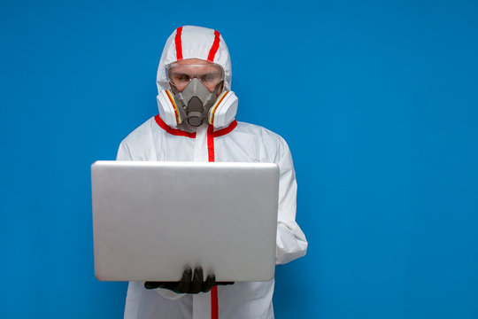 Epidemiologist in a chemical protective suit holds a laptop on a blue isolated background, man, biologist, scientist, virologist types on a computer