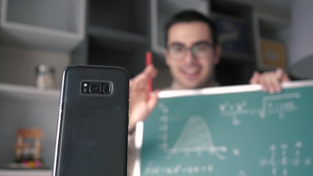 A male Caucasian online educator , holding a chalk board and  adjusting his smartphone mobile device for his video call with his students.